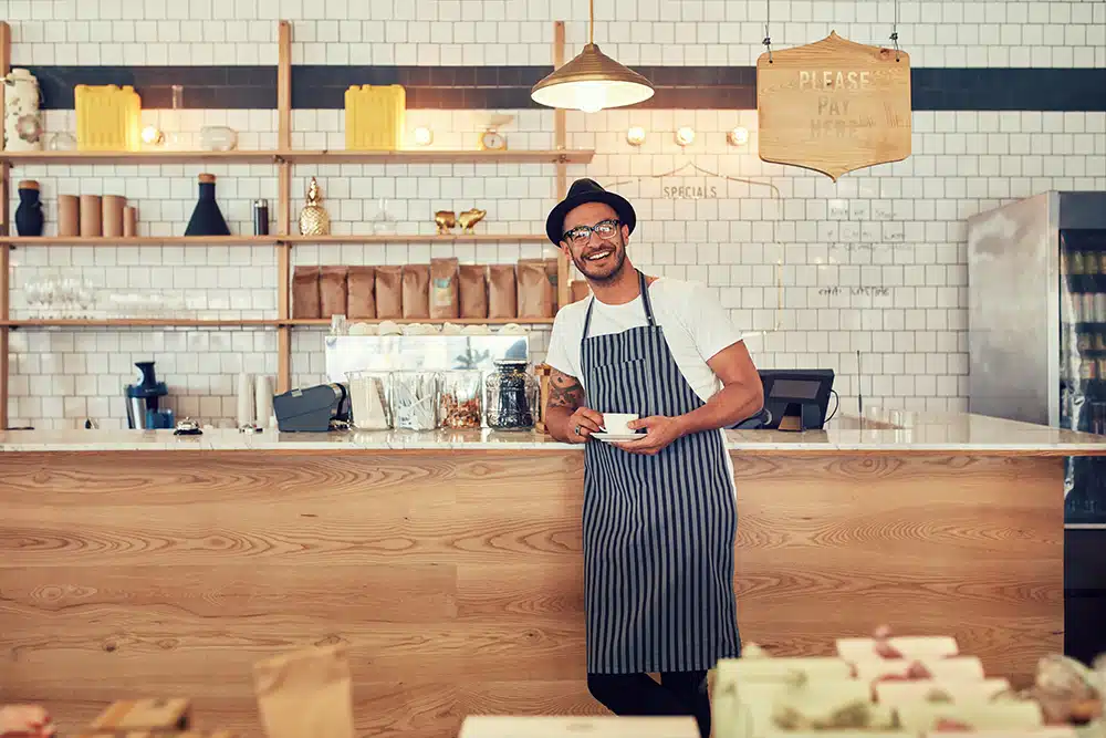 Smiling male barista in apron standing behind a stylish coffee shop counter