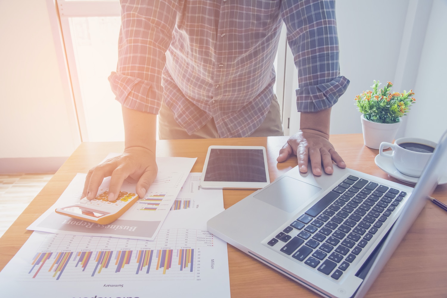 Person calculating financial data at a desk with laptop, tablet, and reports