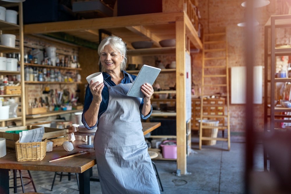 Smiling older woman in an apron holding a ceramic bowl and tablet in a pottery studio