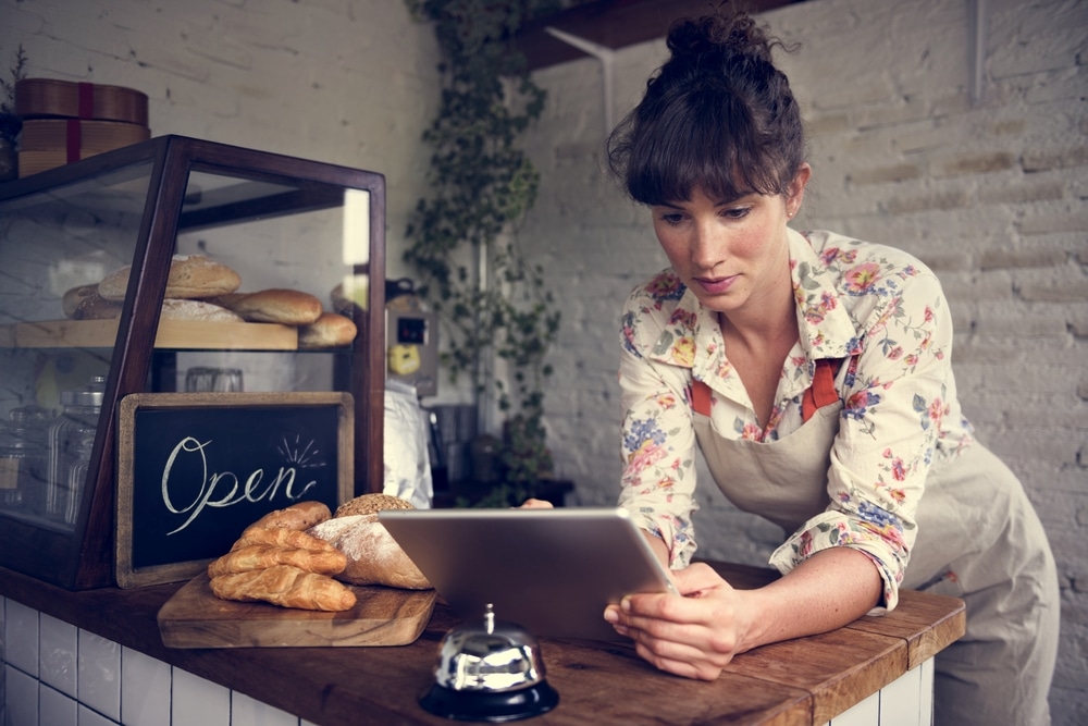 Female bakery owner using tablet behind counter with pastries and open sign