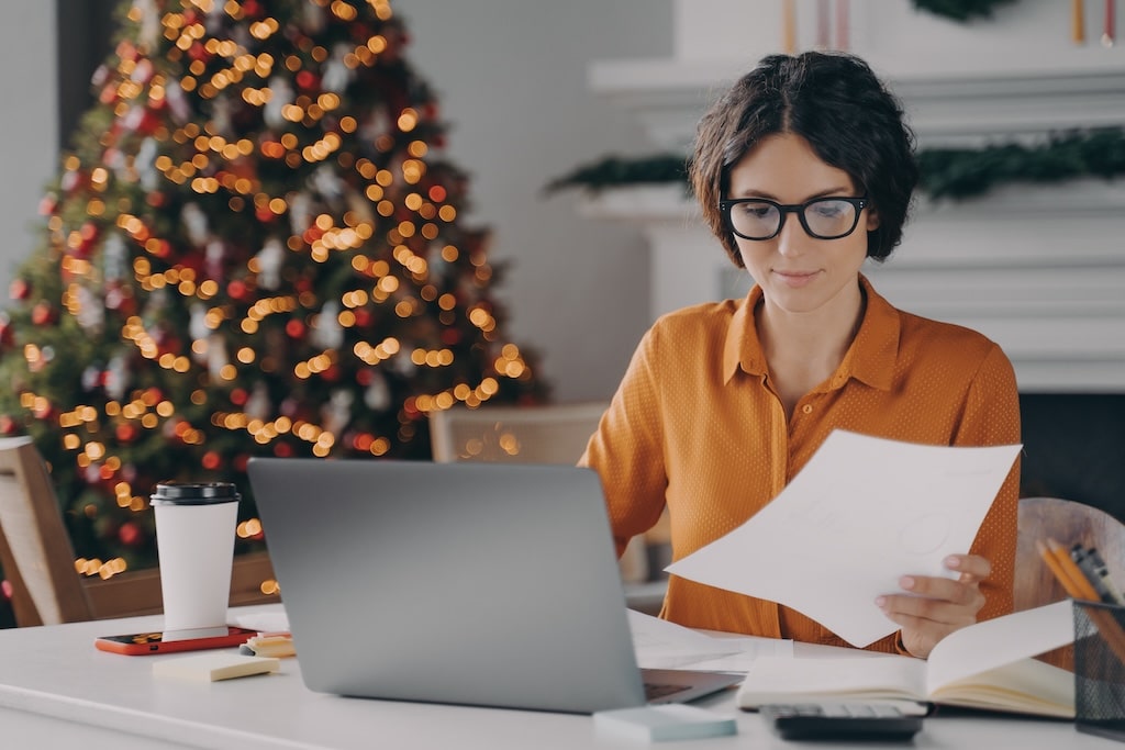 Woman working on laptop while reviewing documents in a festive holiday setting