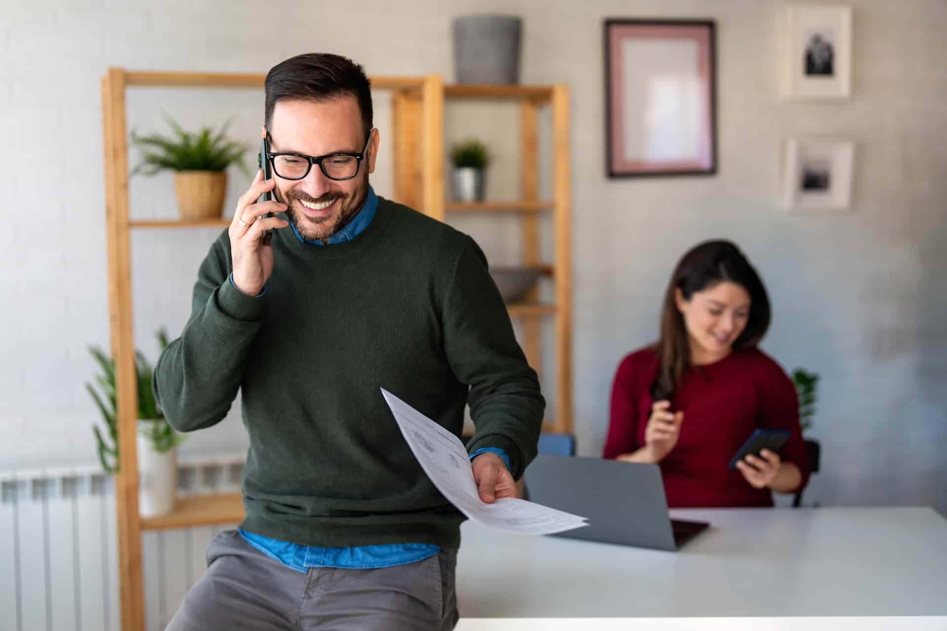 Smiling man on phone holding documents in a modern office with a colleague in the background