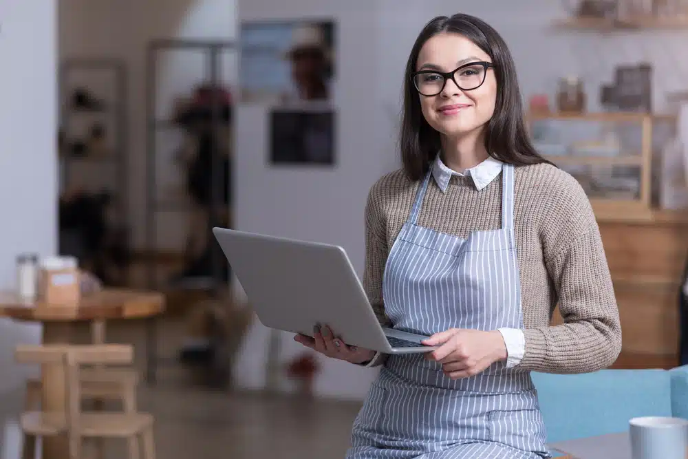 Smiling business owner in striped apron holding laptop inside a modern café or shop