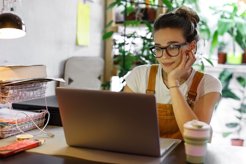 Young woman wearing glasses and overalls working on a laptop at home
